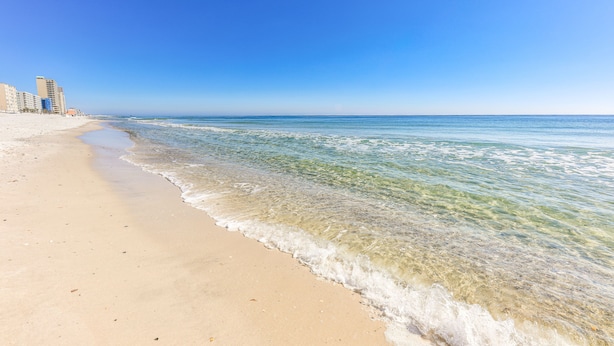Uma praia com águas cristalinas, areal arenoso e edifícios ao fundo.