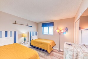 A hotel room with two beds, a nightstand with a lamp, a bedside table, a window with blinds, a floor lamp, and a mirror.