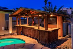 A wooden outdoor bar area with a pool, surrounded by a fence and palm trees.