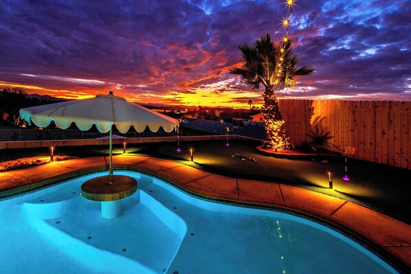 A swimming pool with a fountain, surrounded by a wooden fence, under a sunset sky.