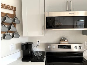 Eine Küche mit einer Edelstahl-Mikrowelle, einer Kaffeemaschine und einem Toaster. Es gibt einen wandmontierten Kaffeebecherhalter und ein Schild mit der Aufschrift 'COFFEE'.
