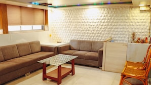 A living room with a sectional sofa, a glass-top coffee table, a wicker chair, and a textured wall. - Hotel Mukesh residency (Pune)