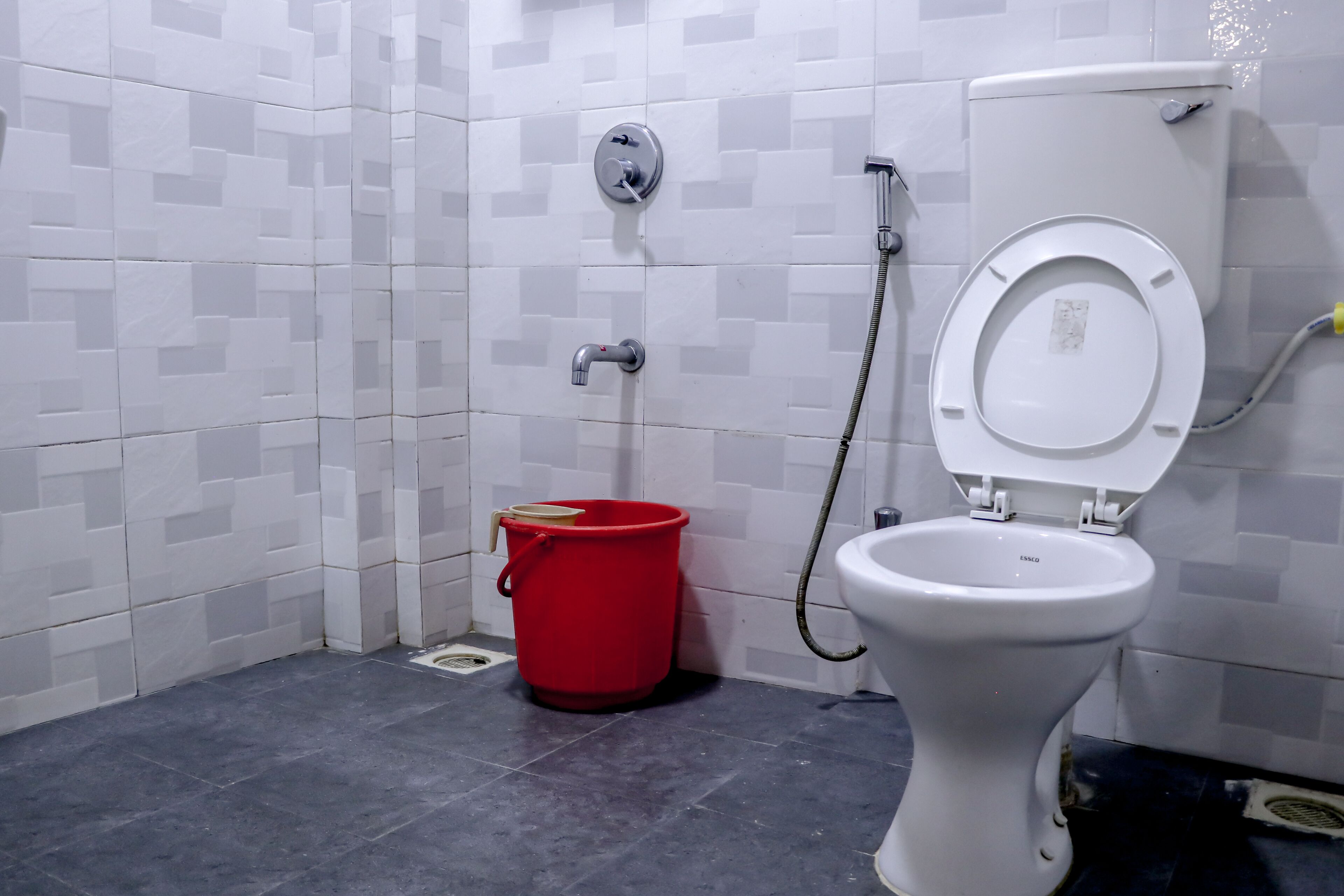 Un baño con un inodoro blanco, un cubo rojo y una manguera de ducha.