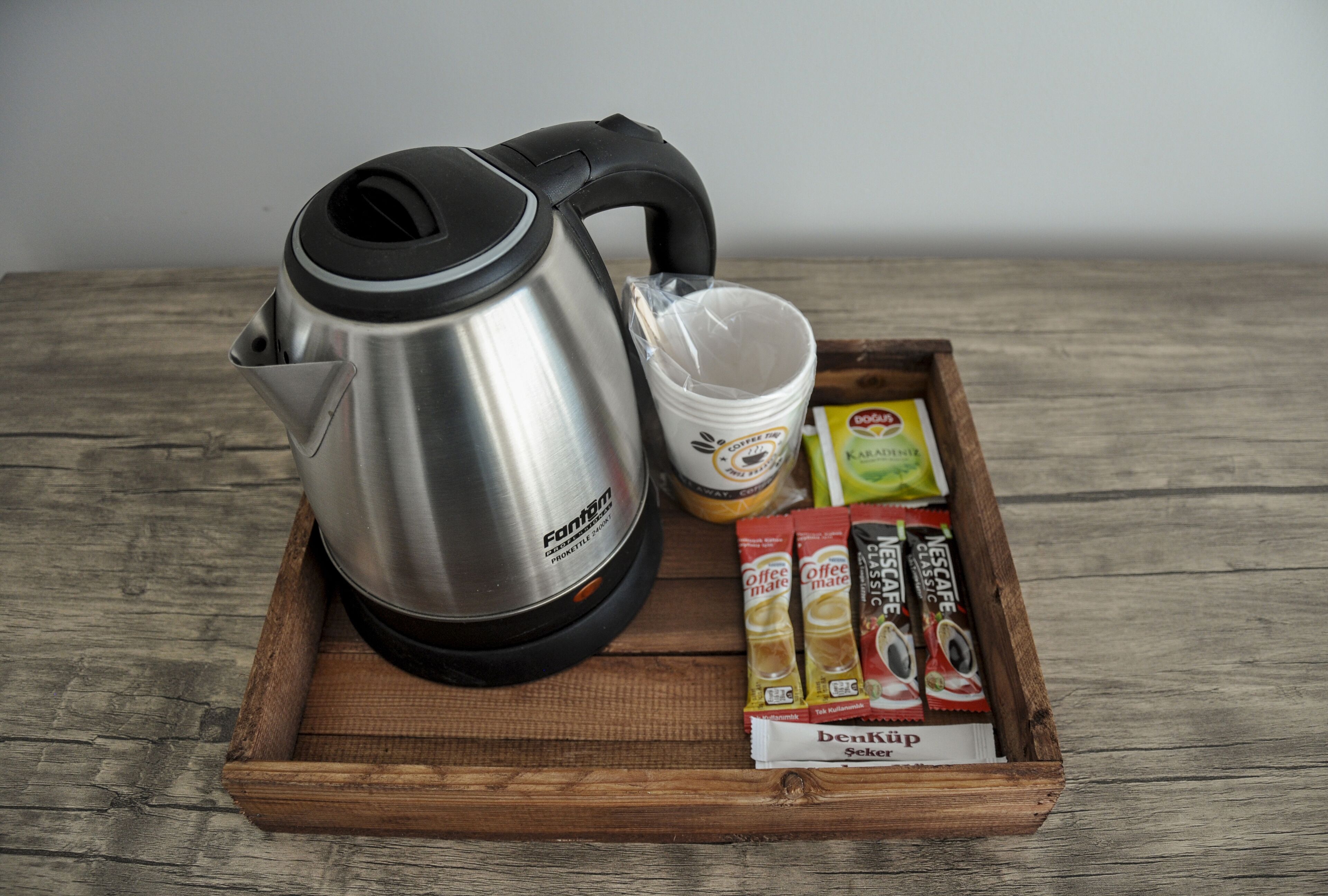 A stainless steel kettle, a plastic cup, and several coffee pods on a wooden tray.