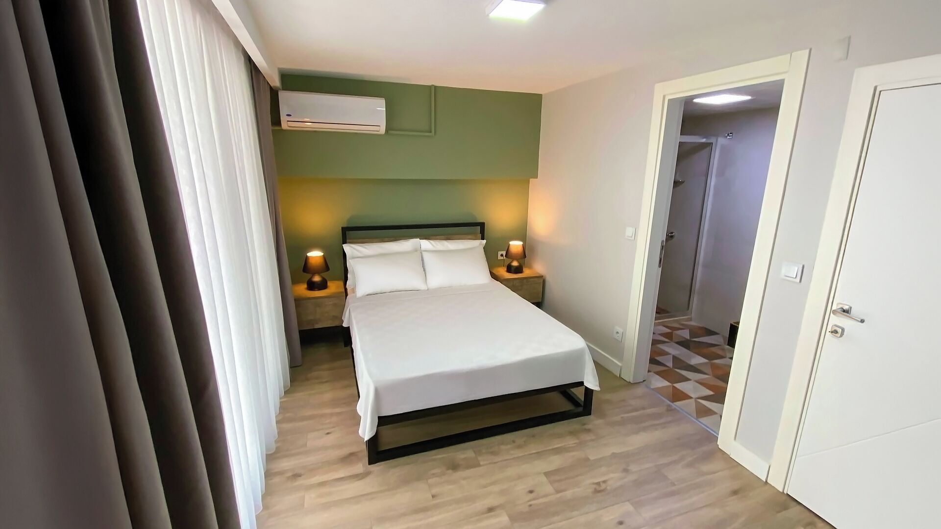 A hotel room with a bed, two bedside tables with lamps, an air conditioning unit, and a door leading to another room.