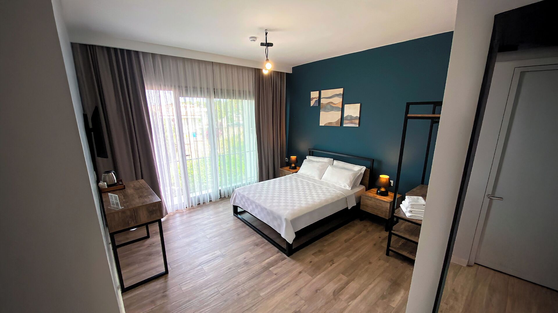 A bedroom with a bed, bedside tables, a desk, and a view of the outdoors through large windows.