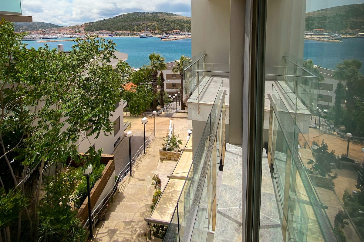 A high-rise building with a glass balcony overlooking a coastal area with boats and a hilly landscape.