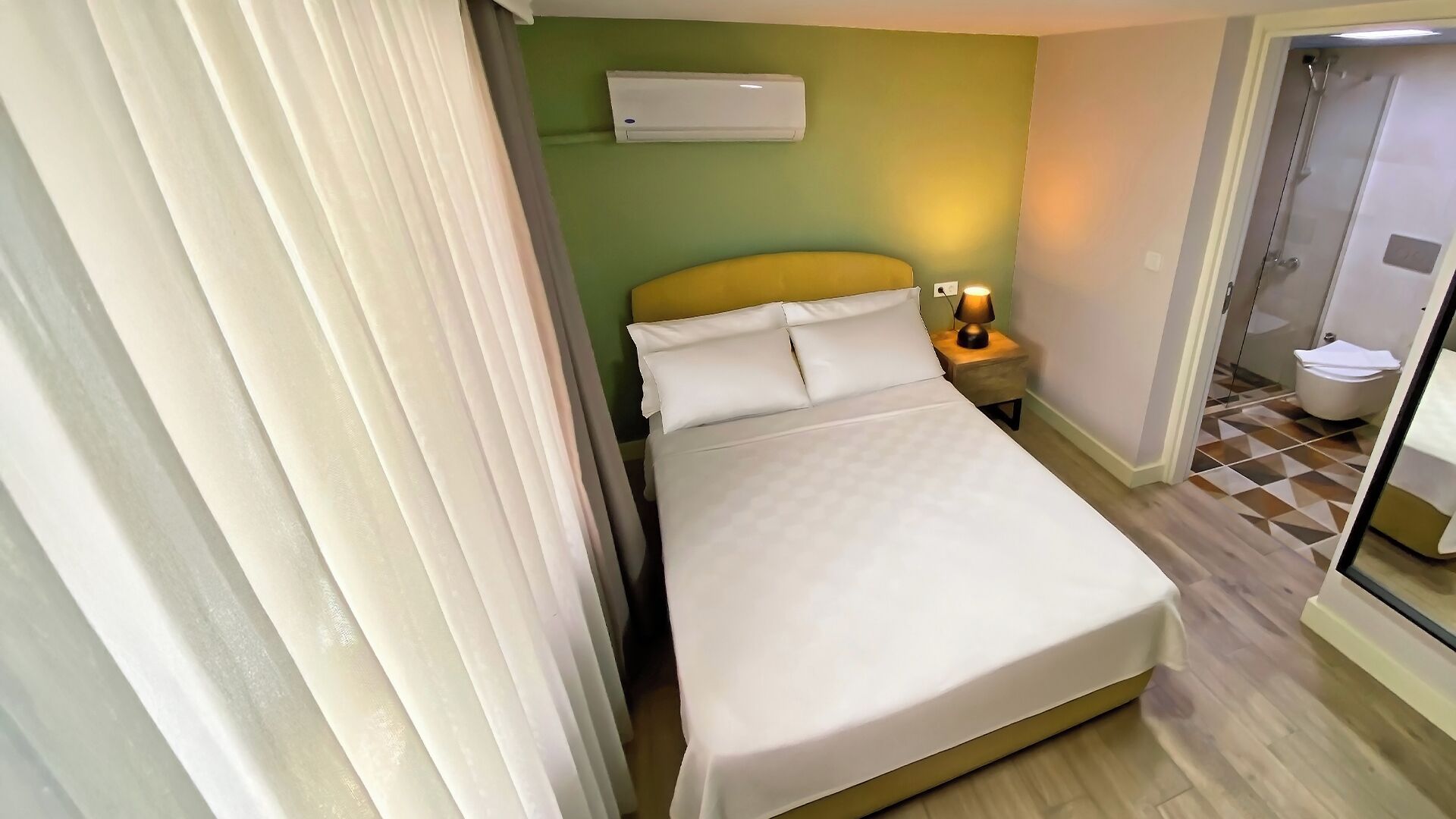 A hotel room with a bed, a nightstand with a lamp, a wall-mounted air conditioner, and a bathroom door.