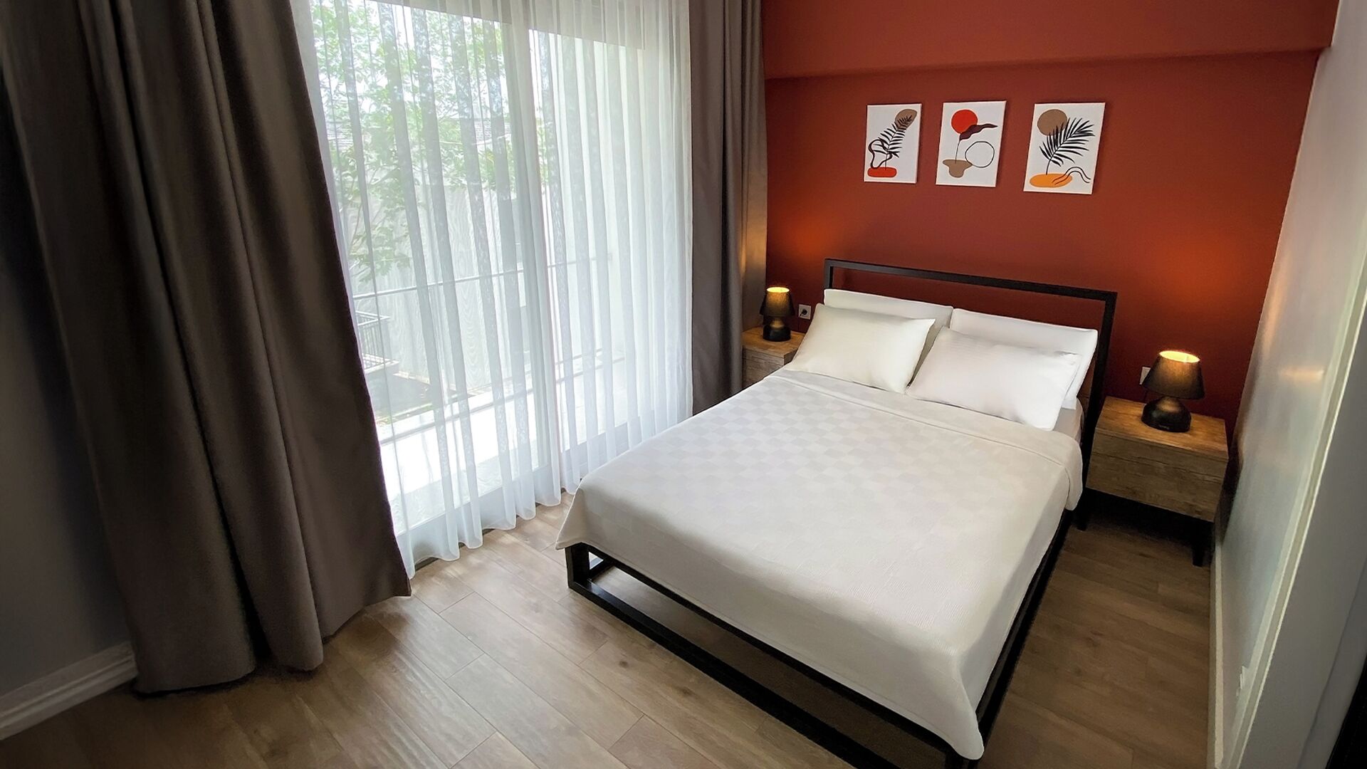 A neatly made bed in a room with a wooden floor, a window with sheer curtains, and three wall-mounted artworks.