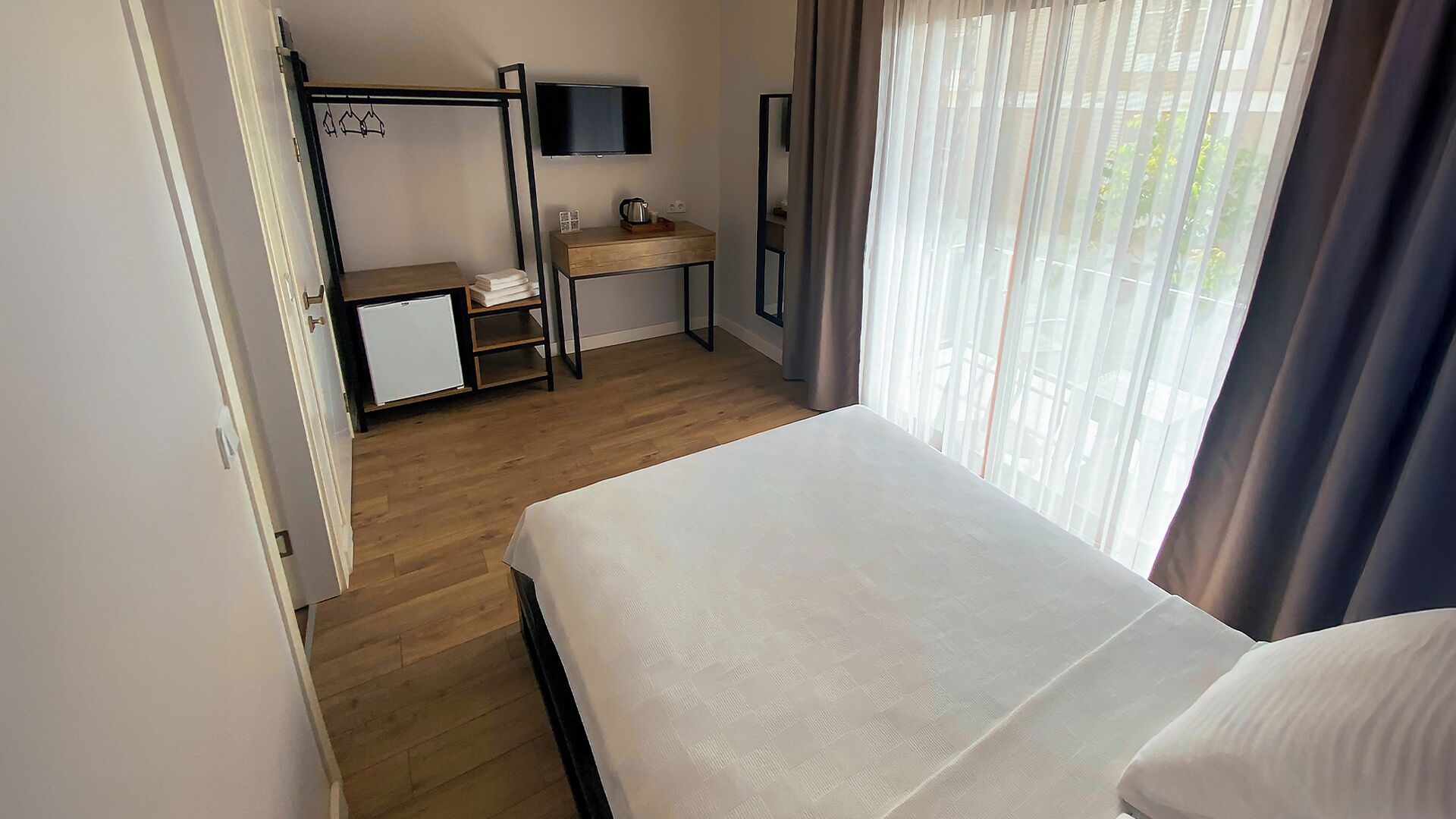 A hotel room with a bed, a desk with a computer, a small fridge, and a TV.
