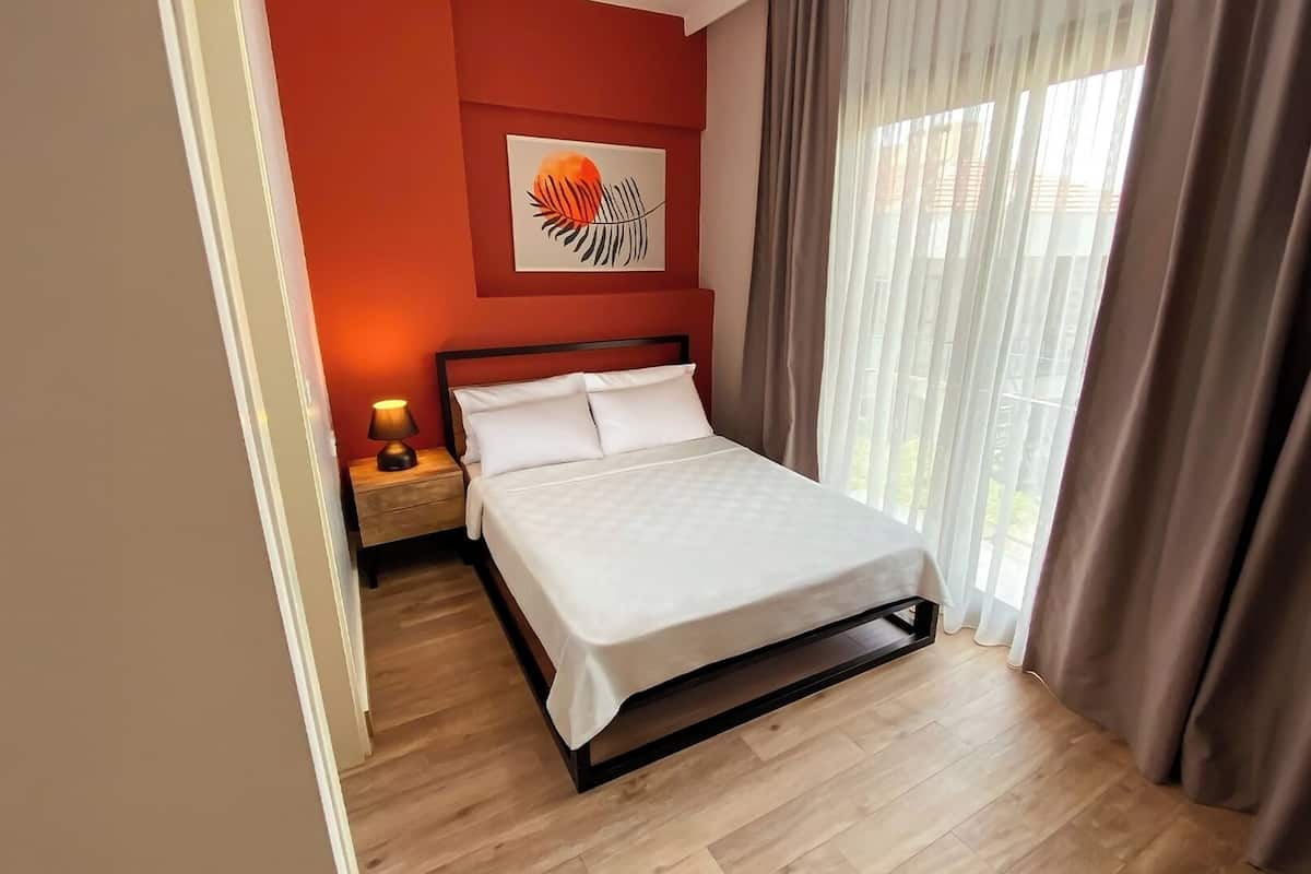 A neatly made bed in a room with a wooden floor, a nightstand with a lamp, and a large window with curtains.
