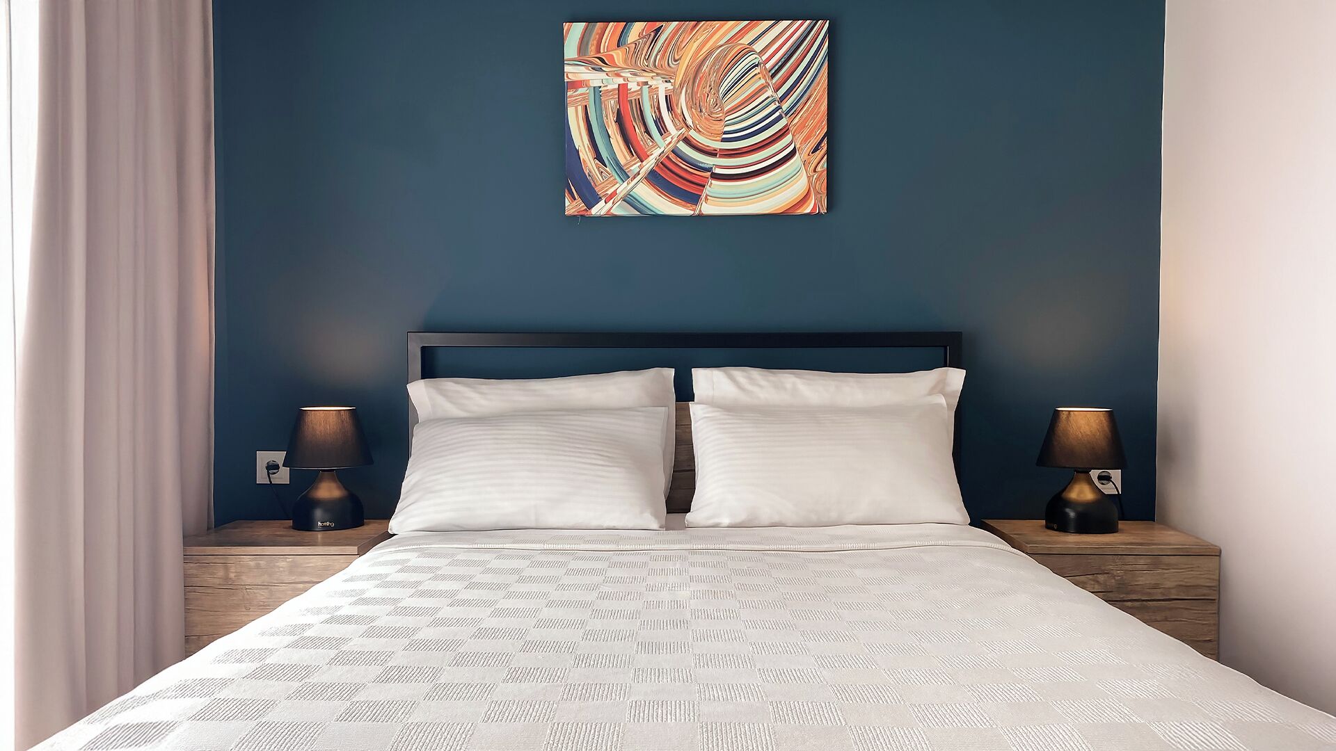 Una camera d'albergo con un letto grande, due comodini con lampade, una parete di colore blu e un quadro astratto dai colori vivaci.