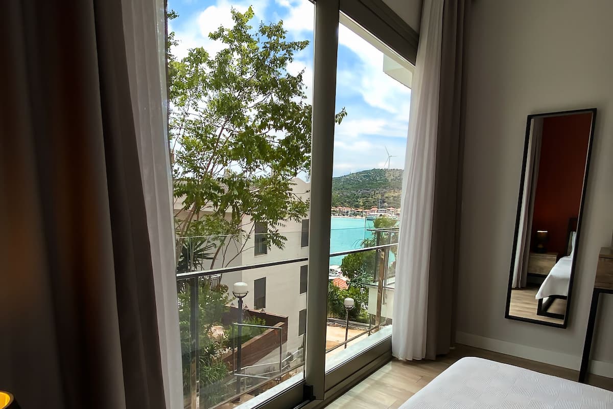 A hotel room with a large bed, a view of a pool and buildings, and a balcony with curtains.