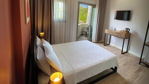 A hotel room with a bed, bedside lamps, a desk with a television, and a view of greenery outside. - Sahil Otel (Cesme)