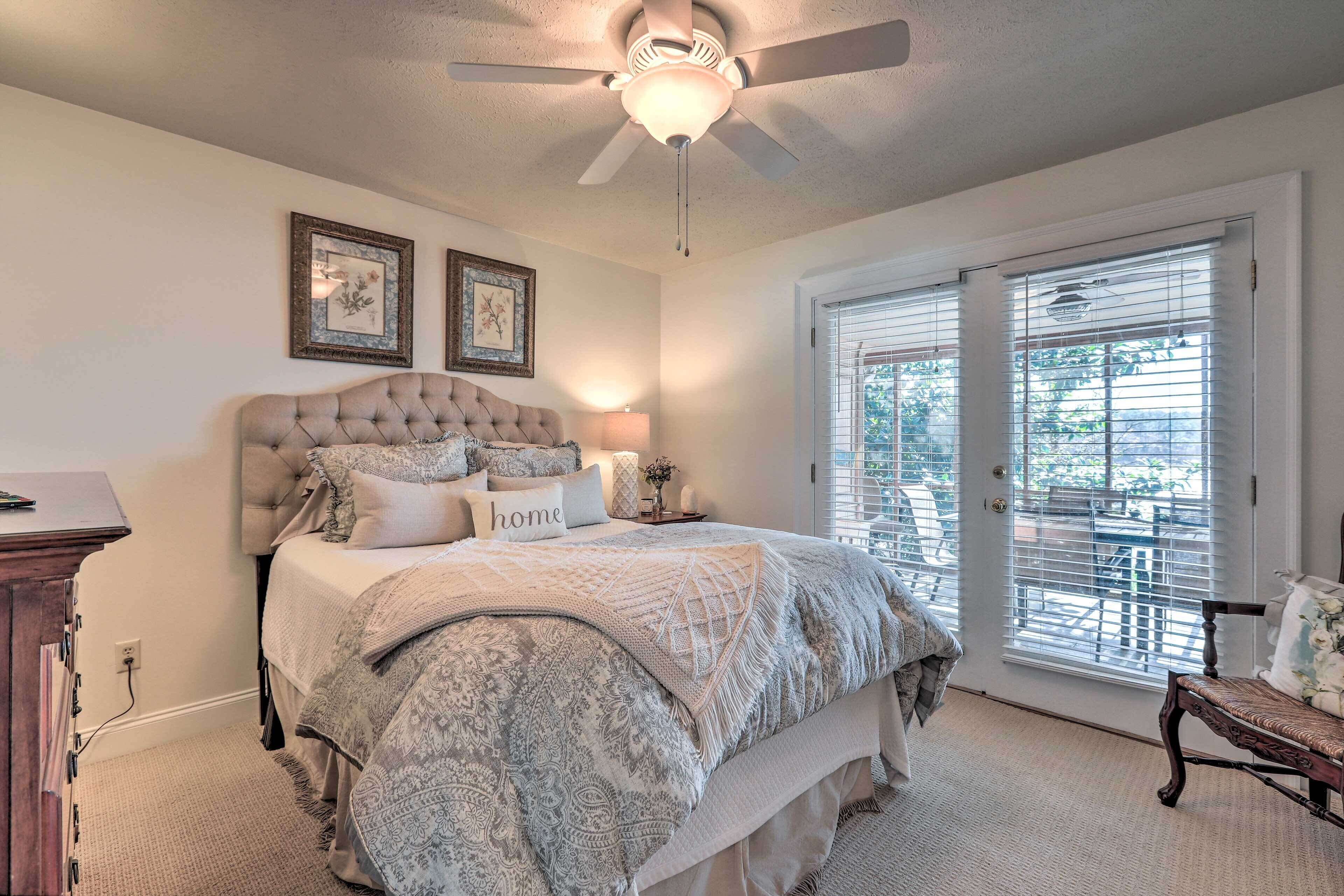 Une chambre à coucher avec une tête de lit capitonnée, un grand lit recouvert d’une literie à motifs, deux tableaux encadrés au mur, un ventilateur de plafond et une vue sur l’extérieur, visible à travers une porte-fenêtre coulissante.
