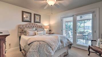 Une chambre à coucher avec une tête de lit capitonnée, un grand lit recouvert d’une literie à motifs, deux tableaux encadrés au mur, un ventilateur de plafond et une vue sur l’extérieur, visible à travers une porte-fenêtre coulissante.