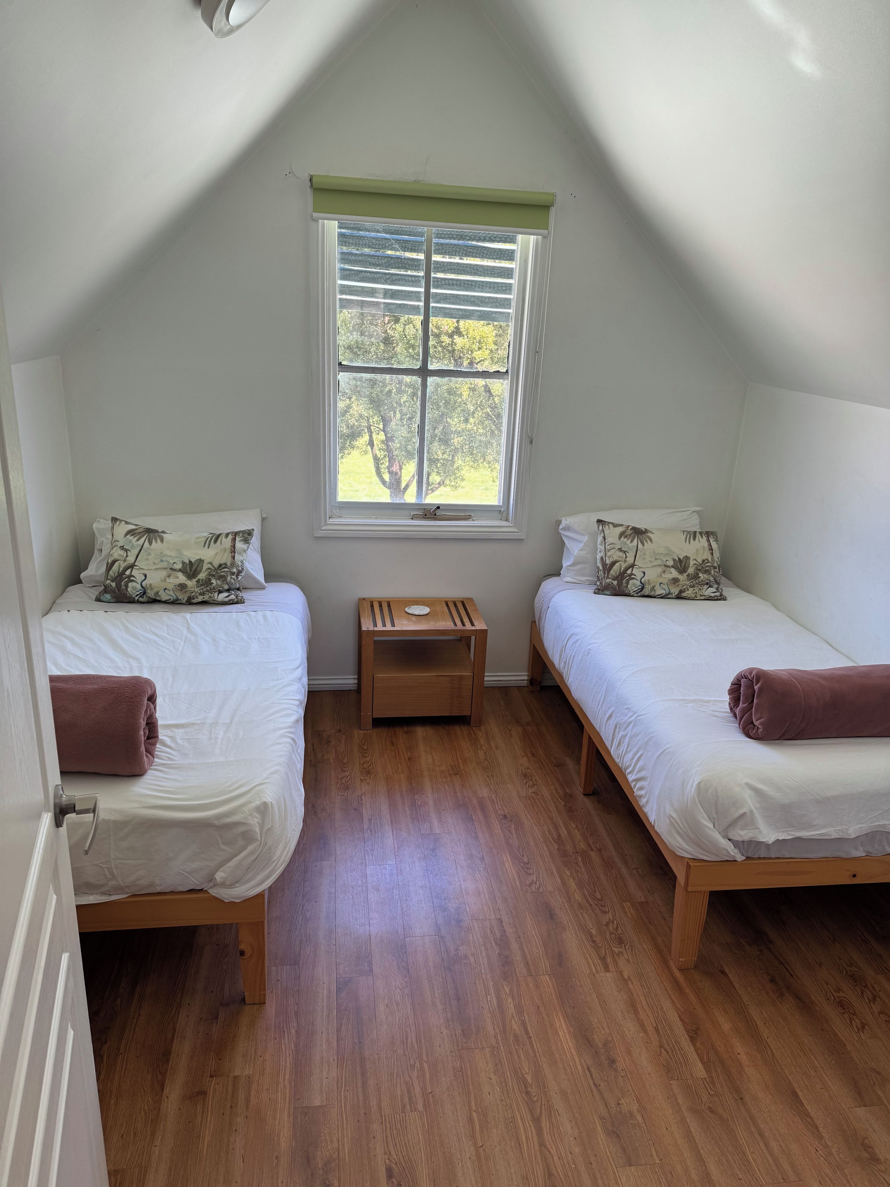 A room with two single beds, a small wooden nightstand, a window with a view of trees, and a sloped ceiling.