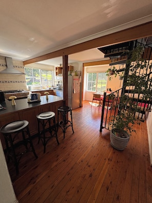 A kitchen with wooden flooring, a central island with stools, a refrigerator, and a window overlooking a garden. - Erica Escape – Where Humans Meet Nature (Erica)