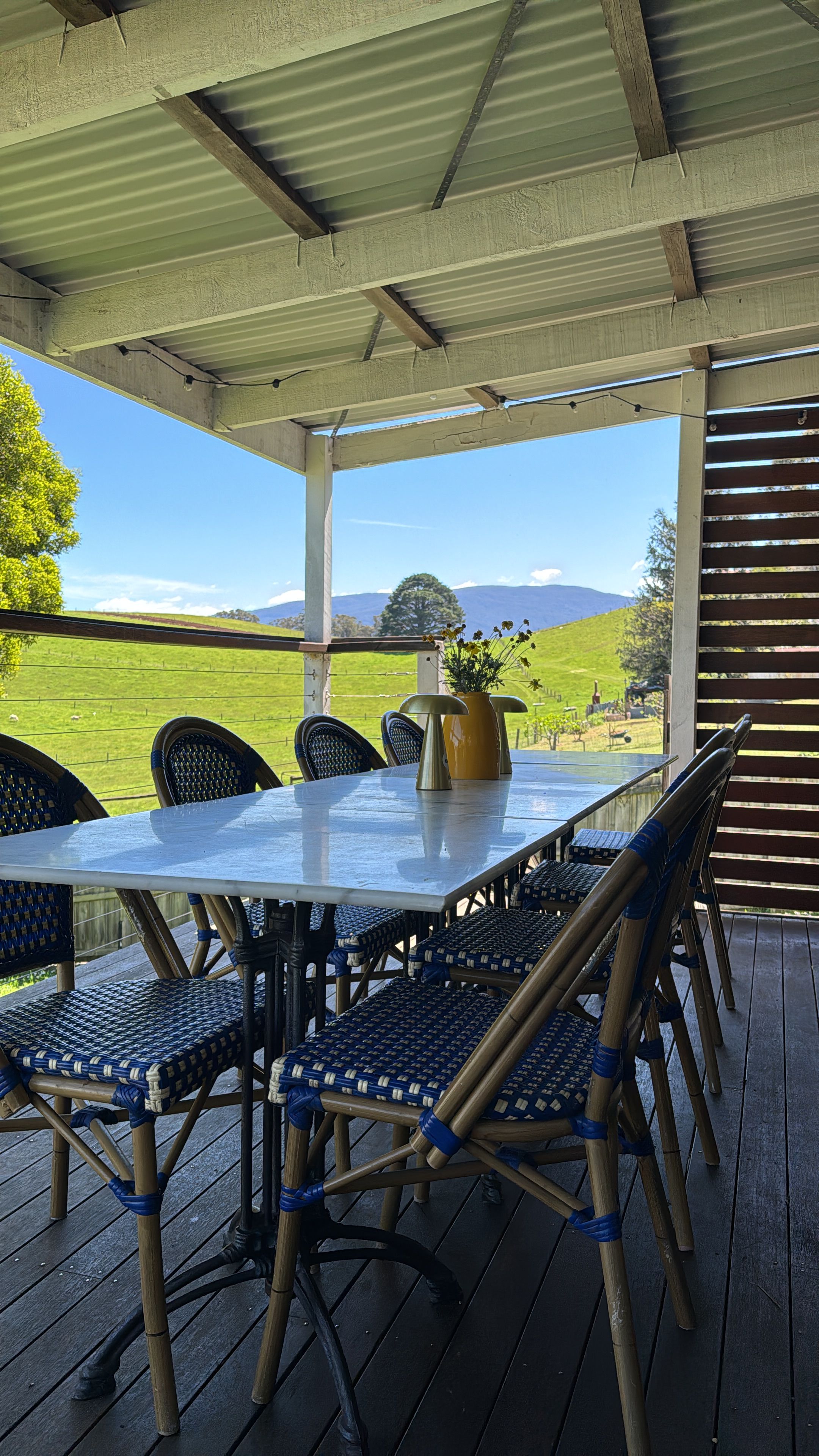 Une terrasse extérieure couverte, avec une table et des chaises, un pot de fleurs et une vue sur un champ verdoyant et des collines.