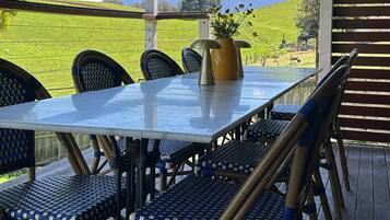 Une terrasse extérieure couverte, avec une table et des chaises, un pot de fleurs et une vue sur un champ verdoyant et des collines.