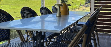 En udendørs overdækket terrasse med et bord og stole, en potteplante samt udsigt over et grønt område og bakker.