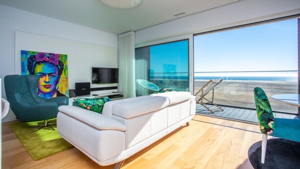 En moderne stue med en hvid sofa, en grøn lænestol, et fjernsyn og et stort vindue med udsigt over stranden.