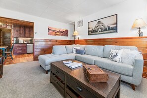 Una sala de estar con un sofá gris, una mesa de centro de madera y una cocina visible a través de una puerta abierta.