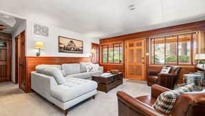 A living room with a grey sofa, a brown leather chair, wooden paneled walls, and a large window with blinds.
