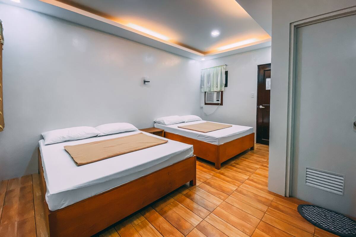 A hotel room with two beds, wooden flooring, a ceiling light, a window with curtains, and a door.