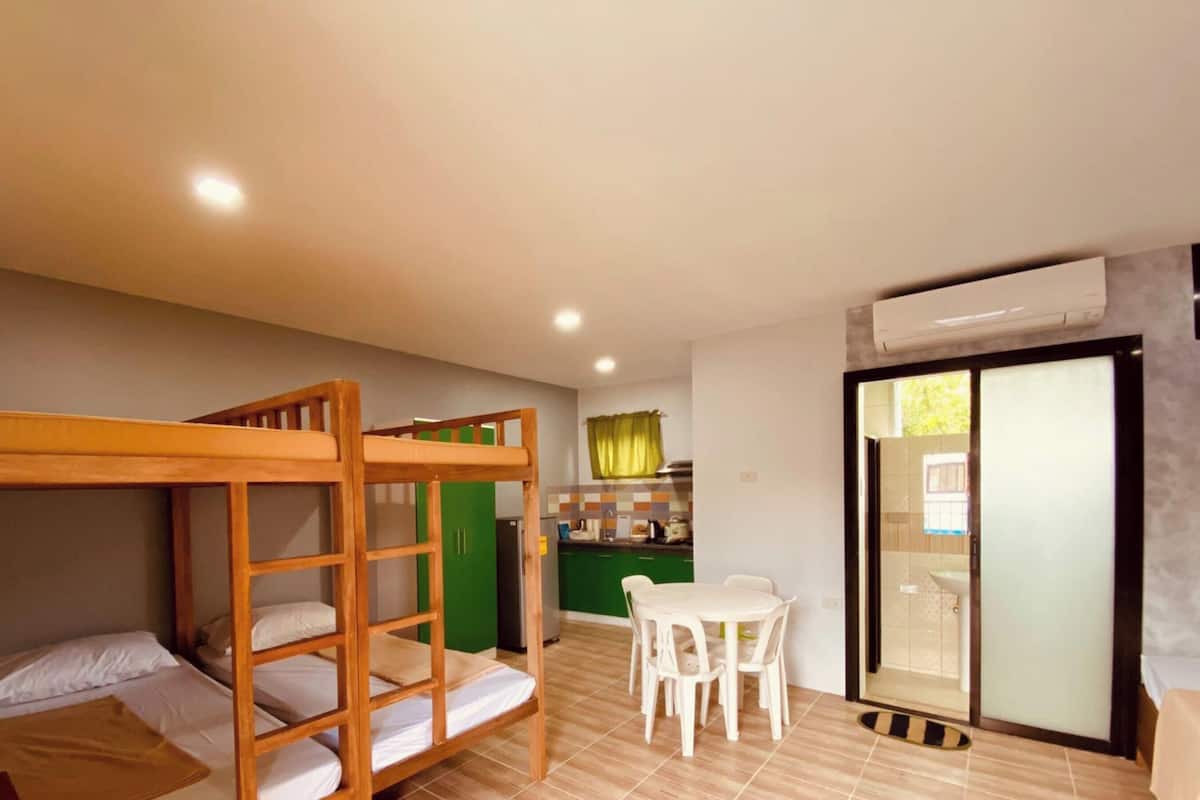 A room with bunk beds, a dining table with chairs, a kitchen area, and a sliding glass door.