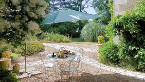 A patio with a table and chairs under a large umbrella, surrounded by lush greenery and a stone pathway.