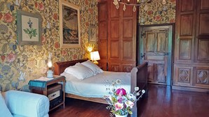 A bedroom with floral wallpaper, a wooden bed, a nightstand with a lamp, a wooden dresser, and a wooden door.