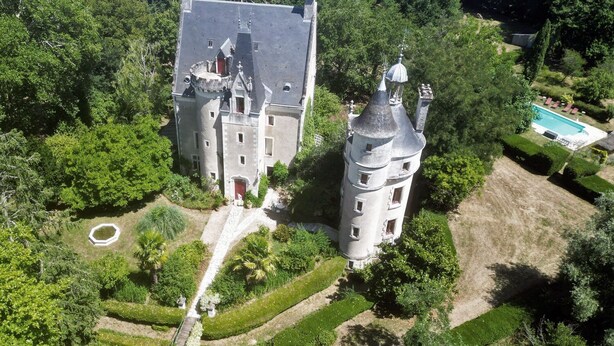 A large, castle-like building surrounded by lush greenery, with a swimming pool and manicured gardens.
