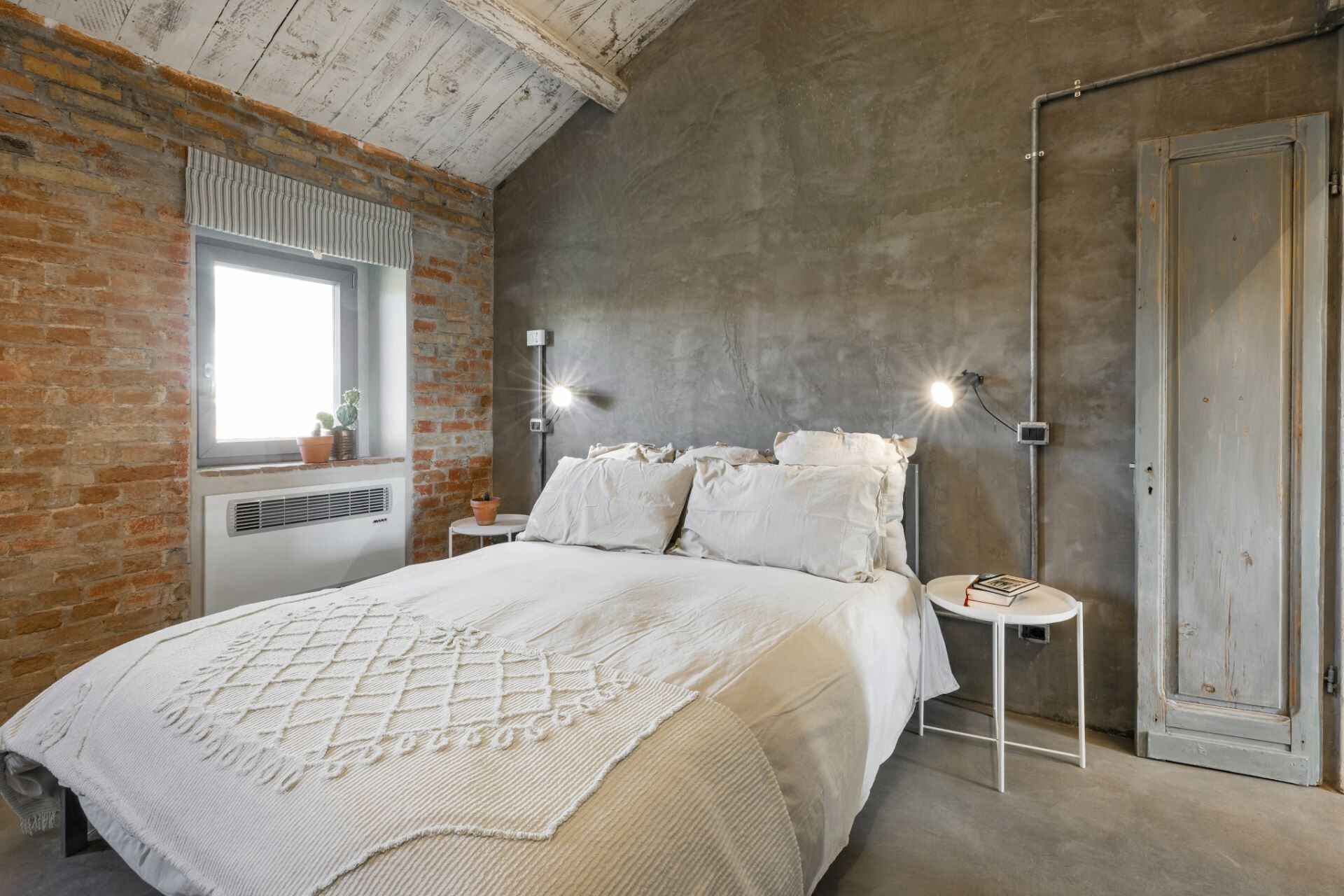 A bedroom with a brick wall, a large window, a bed with white bedding, a bedside table with a lamp, and a door.