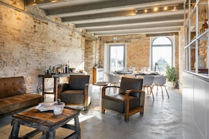 A rustic interior with exposed brick walls, a bar area with bottles, leather seating, and a dining setup with a view through large windows.