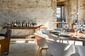 A rustic dining area with a brick wall, a bar with various bottles, and white chairs.