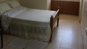 Une chambre à coucher avec un lit, une petite fenêtre avec des rideaux, une table de chevet et un sol carrelé.