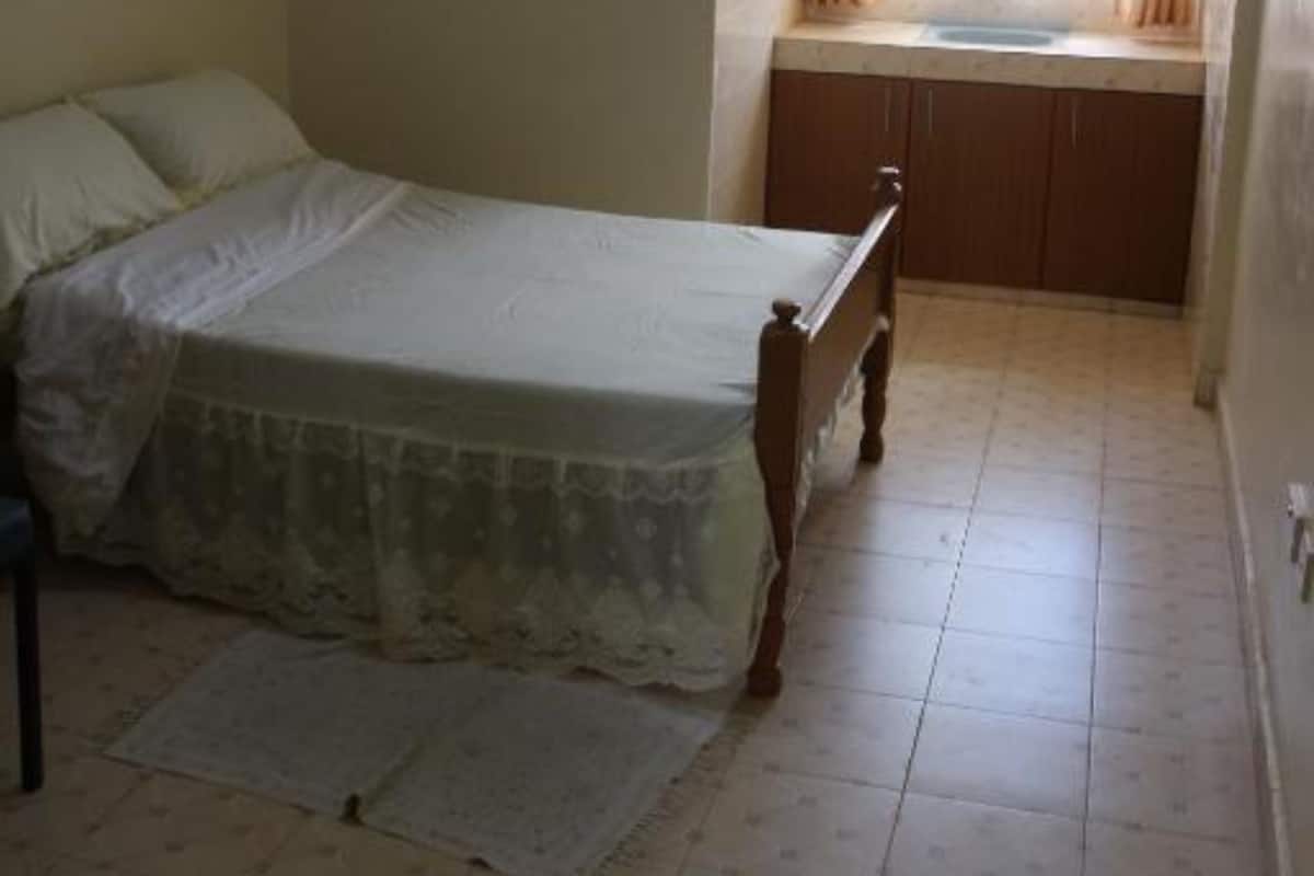 A bedroom with a bed, a small window with curtains, a bedside table, and a tiled floor.