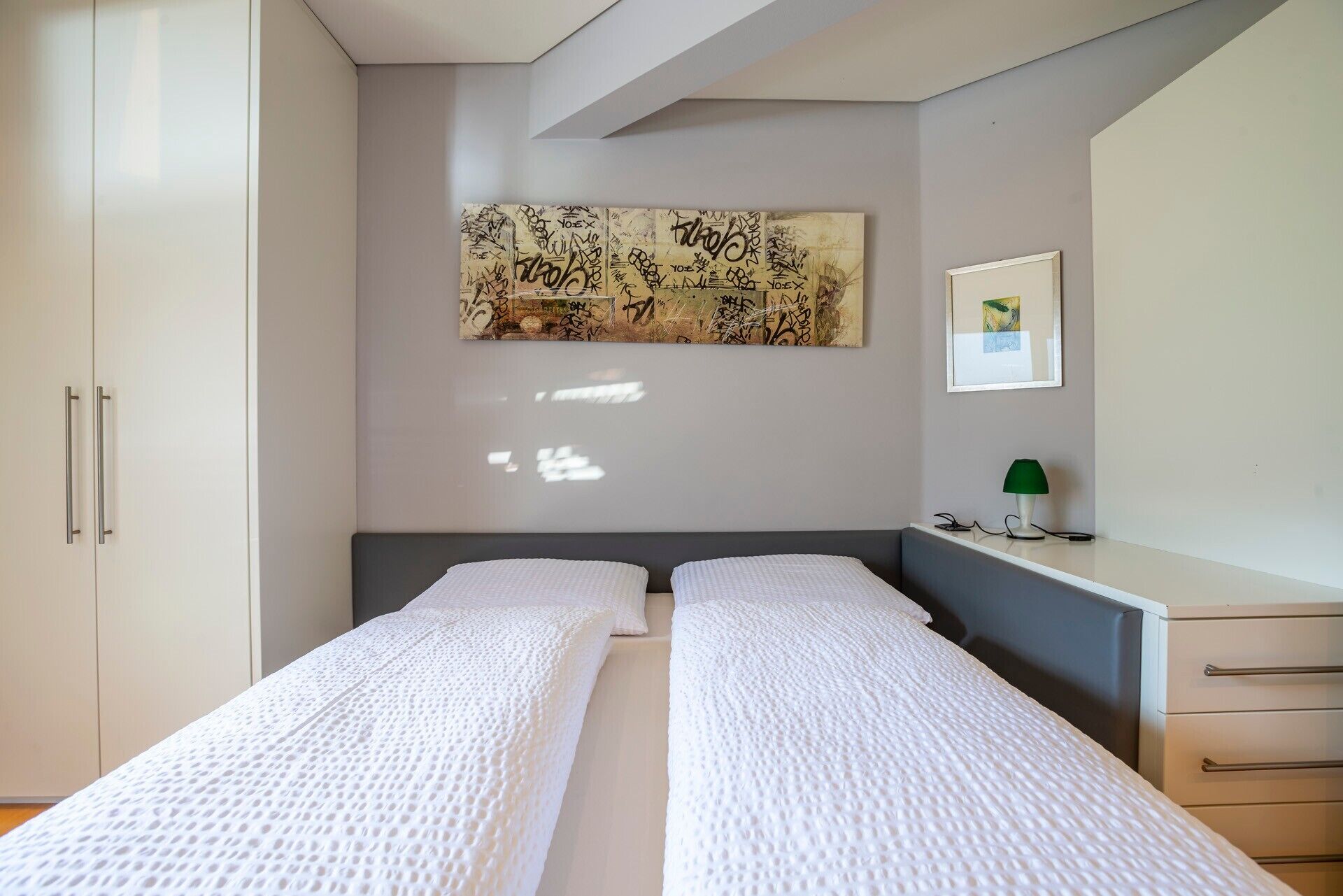 Quarto com duas camas de solteiro, uma pintura abstrata grande, uma fotografia emoldurada, uma mesa de cabeceira com luminária e um guarda-roupa.