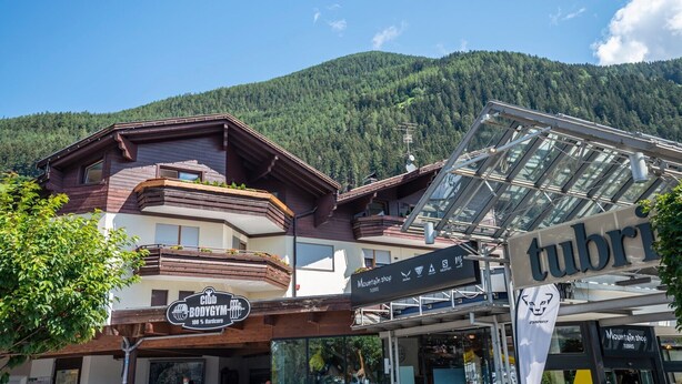 Un magasin de montagne avec une enseigne, une salle de sport appelée Club BodyGym, et une vue sur les montagnes en arrière-plan.