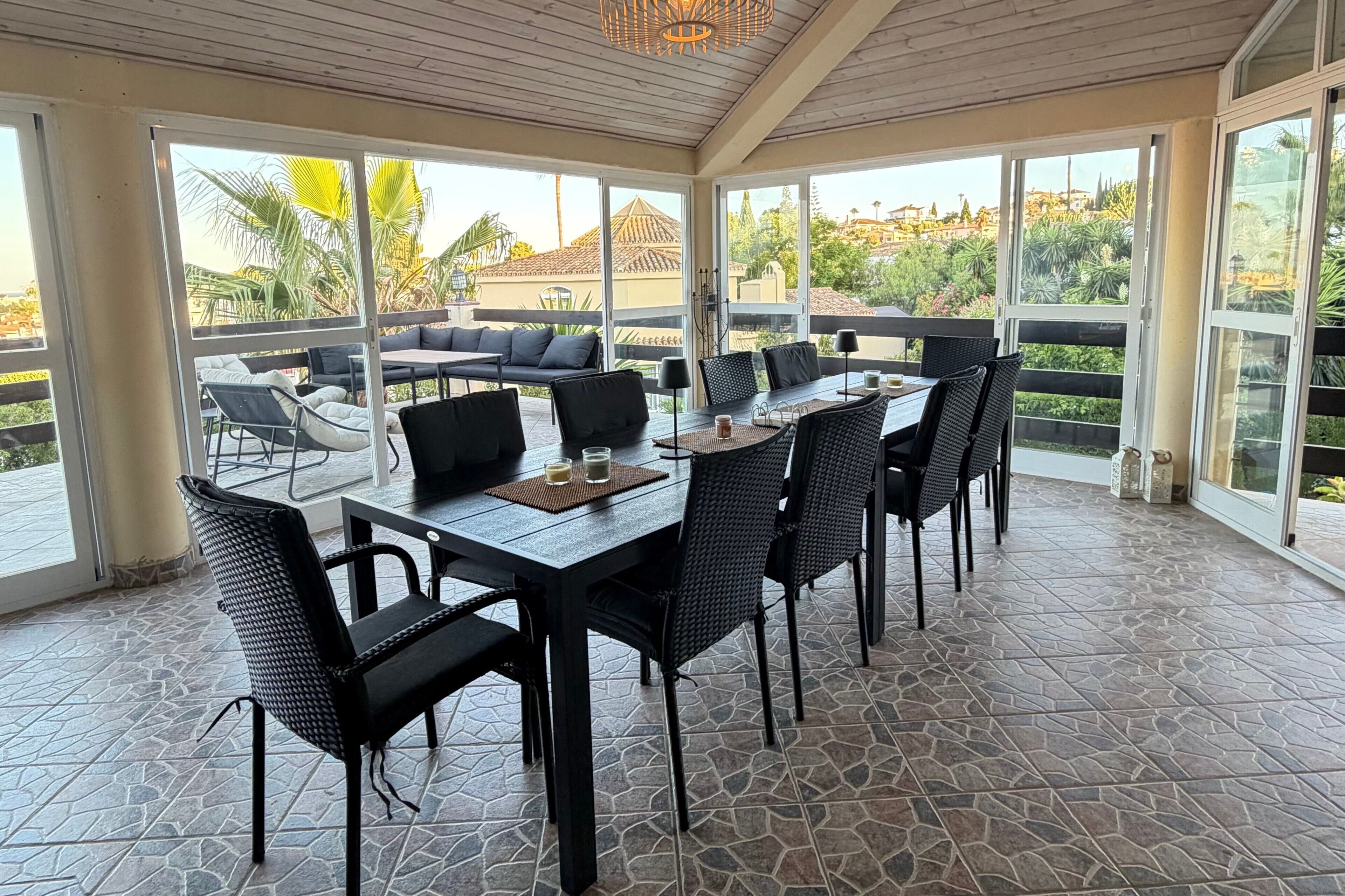 Un área de comedor al aire libre con techo, suelo de losas de piedra, mobiliario negro y amplios ventanales con vistas a una zona residencial.