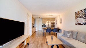 Een moderne woonkamer met een flatscreen televisie, een houten tv-meubel, een bank met decoratieve kussens, een eethoek met stoelen en een keuken op de achtergrond.