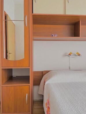 A bedroom with a wooden wardrobe, a bed with a white textured bedspread, and a small shelf with decorative items.