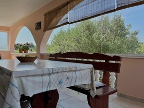 An outdoor dining area with a table and bench, covered by a patterned tablecloth, and a view of trees through a window.