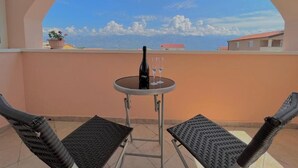 A balcony with two chairs, a small table with a bottle and glasses, and a view of the sea and buildings. - Apartment Nico (Razanac)