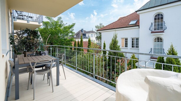 En balkon med et bord og stole, en overdækket sofa og udsigt til omkringliggende huse.