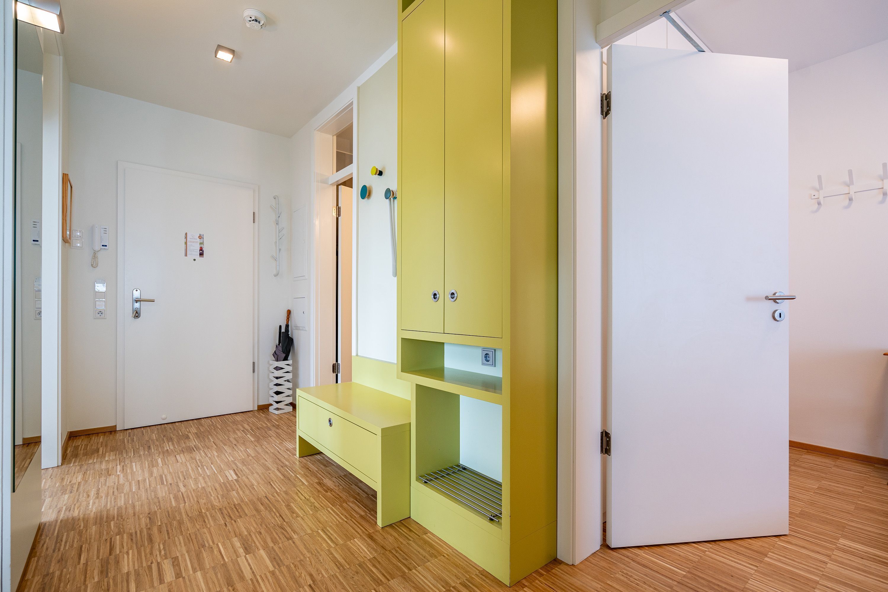 Ein Zimmer mit Holzboden, einem gelben Schrank mit Regalen, einer weißen Tür und einer weißen Wand mit Kleiderhaken.