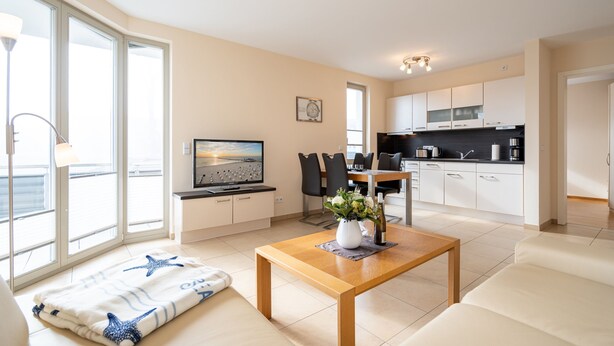 A modern living room with a sofa, a wooden coffee table, and a TV. There is a kitchen area with white cabinets and a dining table. - Villa Estrelia, App. 6 (Heringsdorf)