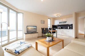 A modern living room with a sofa, a wooden coffee table, and a TV. There is a kitchen area with white cabinets and a dining table. - Villa Estrelia, App. 6 (Heringsdorf)