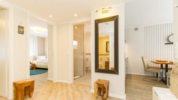 Ein Hotelzimmer mit Holzboden, einem großen Spiegel, einem Essbereich mit Tisch und Stühlen sowie einem durchgehenden Türspalt sichtbaren separaten Badezimmer.